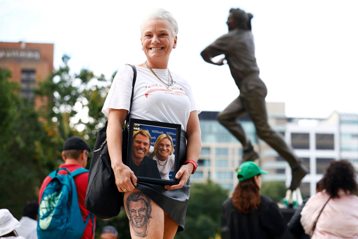 A member of the public with a Shane Warne tattoo poses in front of a statue of Shane Warne before the state memorial service