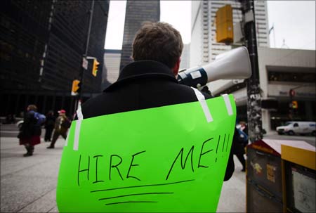 A man, with a sign strapped to his back, uses a megaphone to attract the attention of potential employers.