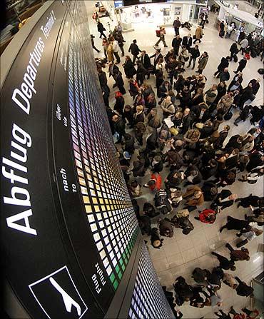 Passengers stand under a flight information display board at Munich Airport.