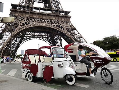 A velo taxi (R), or bicycle taxi, drives past a tuk-tuk, or three wheeled auto rickshaw, near the Eiffel tower in Paris.