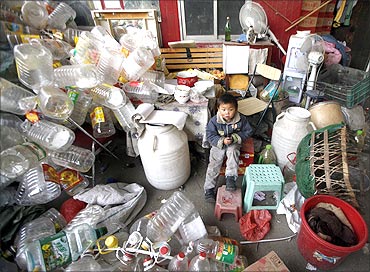 A boy sits outside the front of a small shelter next to a pile of rubbish at a recycling centre.
