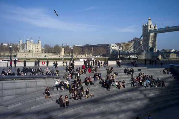 People sit on the banks of the River Thames in front of Tower Bridge.