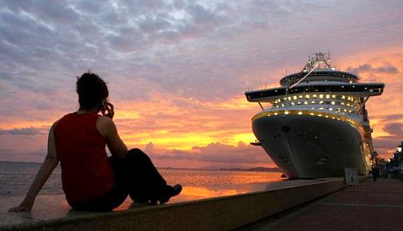 A woman talks on her phone while watching the sun set behind a cruise ship.