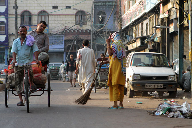 A woman sweeps a street in Old Delhi as a rickshaw driver transports a passenger.