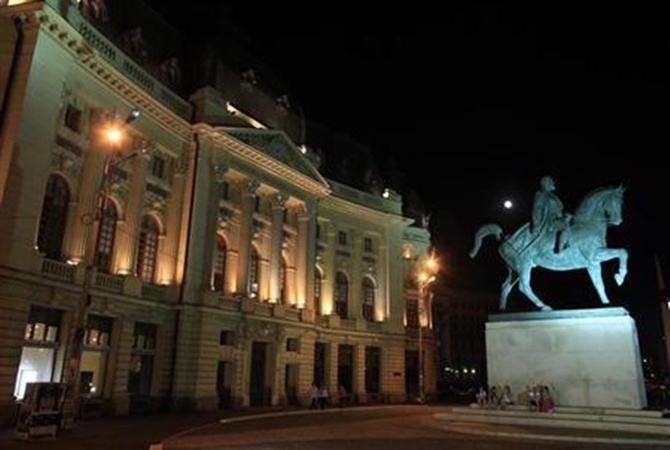 People sit near a statue of King Carol I, the founder of Romania's royal dynasty, as the moon rises in Bucharest.