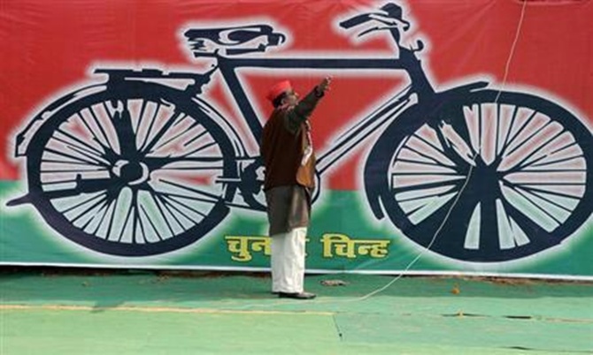 A Samajwadi Party worker gestures in front of a banner with the party's electoral symbol.