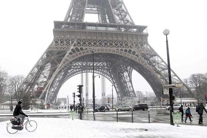 A man rides his bicycle as he makes his way along a snow-covered sidewalk near the Eiffel Tower.