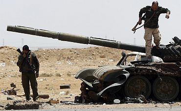Rebel fighters walk near a destroyed tank near the front line along the western entrance gate of Ajdabiyah