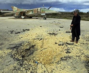 A man shows a crater caused by what he says is the impact of a missile fired from a Libyan army aircraft as he stands in front of an old military plane at a military airport runway in the eastern Libyan town of Al Abrak