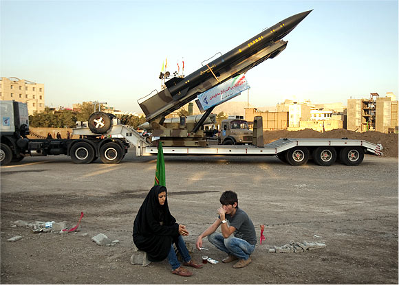 A couple rests as they sit in front of an Iranian-made Zelzal missile at a war exhibition