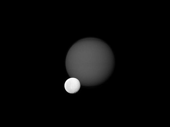 The Cassini spacecraft views the cratered surface of Saturn's moon Tethys in front of the hazy orb of the planet's largest moon, Titan
