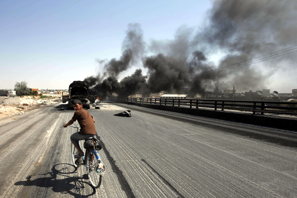 A Syrian boy rides a bicycle as smoke rises over the Syrian city of Aleppo after missiles fired from a fighter jet hit petrol tankers in the Bab al-Nayrab district