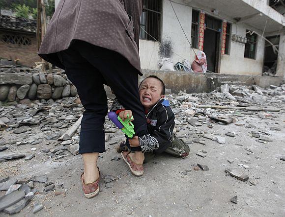 A boy holds his mother's leg as he cries in front of their damaged house