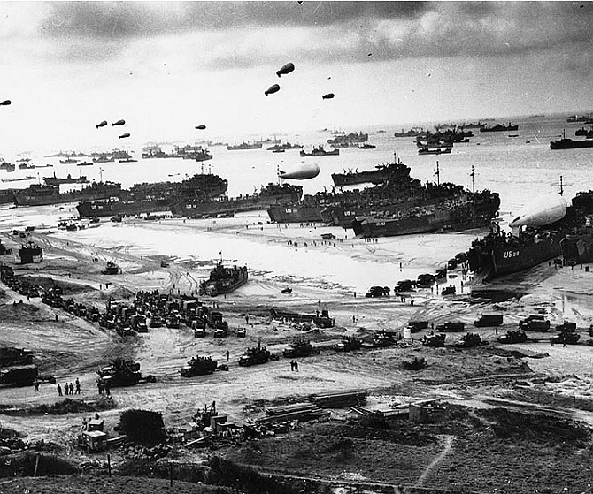 Landing ships putting cargo ashore on one of the invasion beaches, at low tide during the first days of the operation, June 1944. Among identifiable ships present are USS LST-532 (in the center of the view); USS LST-262 (3rd LST from right); USS LST-310 (2nd LST from right); USS LST-533 (partially visible at far right); and USS LST-524. Note barrage balloons overhead and Army half-track convoy forming up on the beach.
