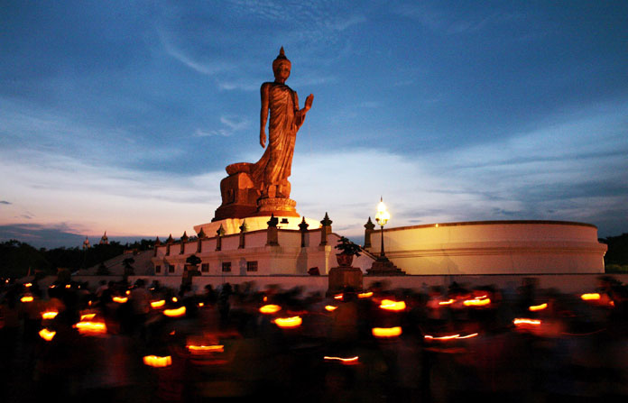 People walk around the Buddha statue at sunset holding candles for prayers at Buddhist Park on Visakha Puja Day in Nakhon Pathom