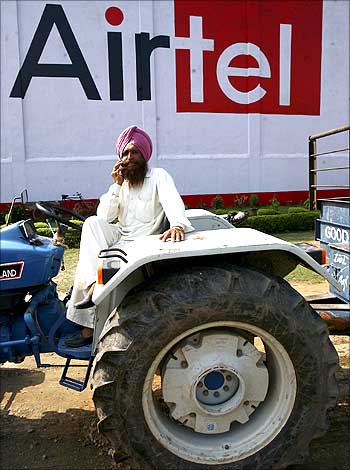 A tractor driver talks on a mobile phone in front of an advisement of Bharti Airtel.