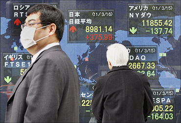 A man wearing a mask walks past an electronic board displaying movements in various market indices around the world, in Tokyo.