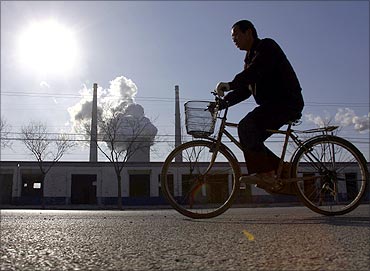 A chimney billows smoke from a coal-burning power station behind a workman riding a bicycle.