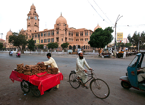 A man sells dry figs from his cart as another rides past on a bicycle in front of the Karachi Municipal Corporation (KMC) building.