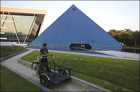 A man pushing a tricycle cart walks in front of a pyramid-shaped building made out of glass in the Infosys campus at Electronics City in Bangalore.