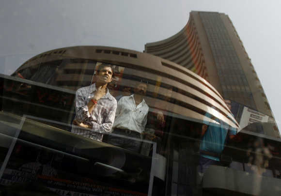 BSE building is reflected on a glass window as people look at a large screen displaying India's benchmark share index in Mumbai.