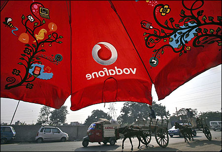 Men ride their horse carts past an umbrella with a Vodafone logo on a road in Jammu.