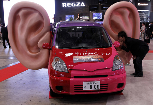 A man talks into a large model of a ear which has recording capabilities on a car.