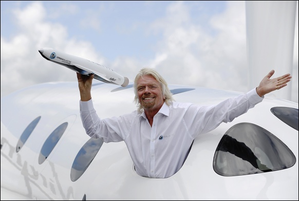 Richard Branson waves a model of the LauncherOne cargo spacecraft from a window of an actual size model of SpaceShipTwo on display