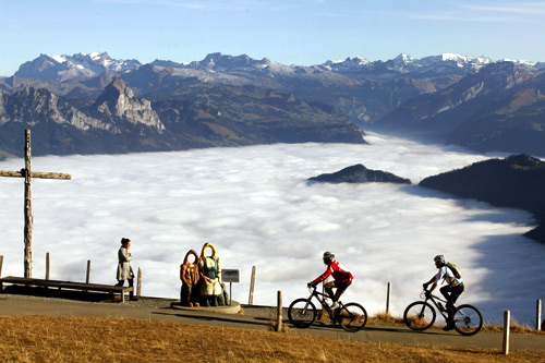 The valleys behind are covered by clouds as bikers ride during sunny autumn weather below the peak of Mount Rigi , at 1,797 m (5,896 ft) above sea level, near Lake Lucerne.