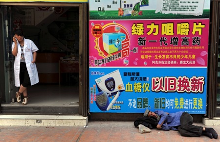 Ethnic Uighur man sleeps in front of posters advertising medical products as an employee talks on her mobile phone at the entrance of a pharmacy in Aksu.