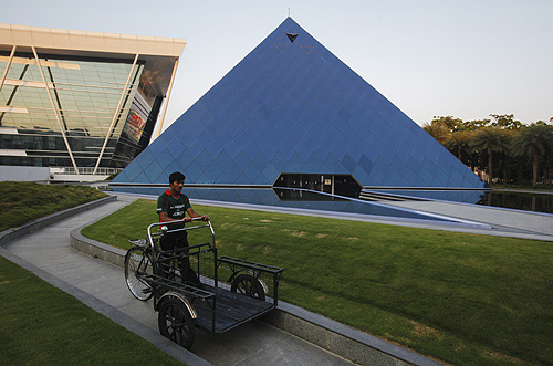 A man pushing a tricycle cart walks in front of a pyramid-shaped building made out of glass in the Infosys campus at Electronics City in Bangalore.