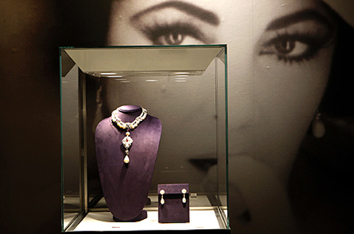 A photograph of Elizabeth Taylor is seen behind jewels on display.