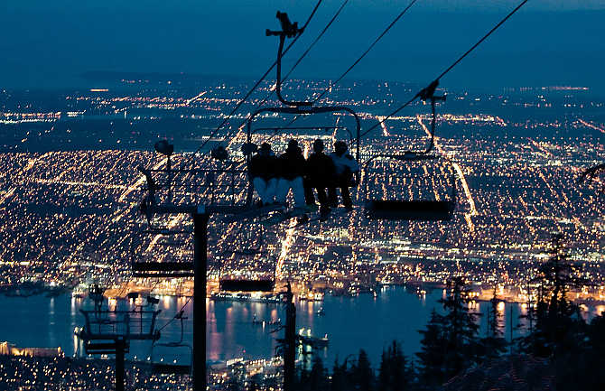 Snowboarders ride a chair lift on one of the many runs during night skiing on Grouse Mountain with the city of Vancouver, British Columbia down below, in Canada.