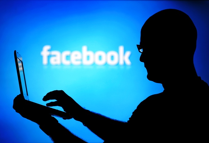 A man is silhouetted against a video screen with an Facebook logo.