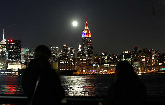 A full moon rises behind the Empire State Building and the skyline of New York as people watch from a park along the Hudson River in Hoboken, New Jersey.