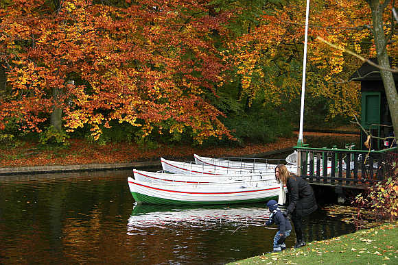 A mother and her child play near the water in front of boats and autumn trees in Frederiksberg Garden in Copenhagen.