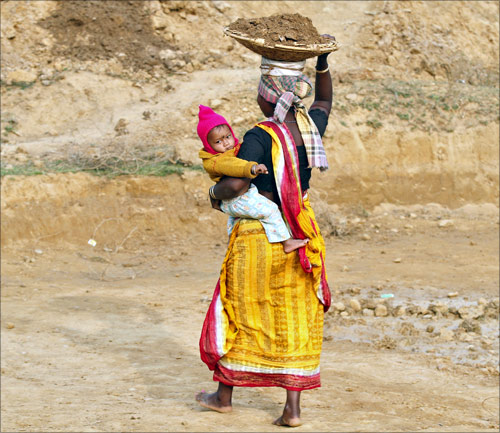 A village woman holds her child while carrying clay on her head as she works at a road construction site under National Rural Employment Guarantee Act.