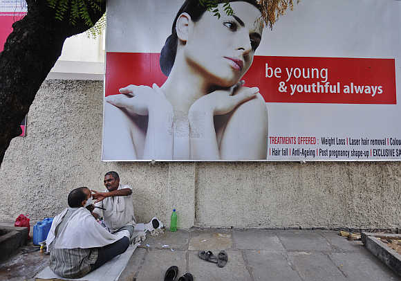A man receives a shave from a street side barber in front of an advertisement for beauty treatments in Hyderabad.