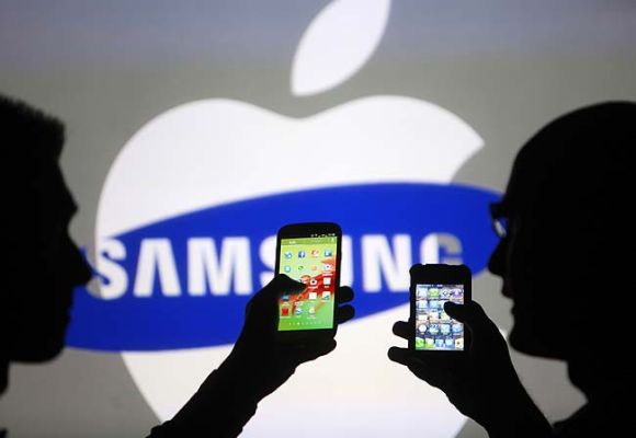 Men are silhouetted against a video screen as they pose with Samsung Galaxy S3 and iPhone 4 smartphones in this photo.