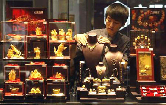 An employee adjusts a gold necklace on a displaying model near glass cases containing figurines at a shop in Wuhan, Hubei province.