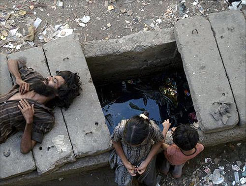 A man sleeps on a gutter cover as children play in a slum in Mumbai.
