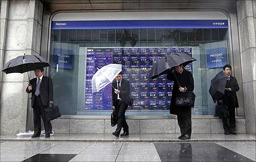 Pedestrians holding umbrellas stand in front of a stock index board showing various stock prices outside a brokerage in Tokyo.