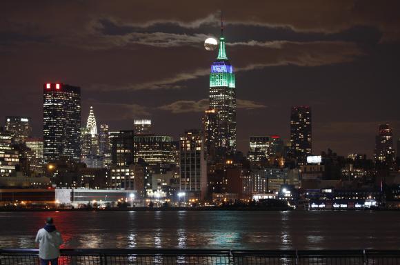 A full moon rises behind the Empire State Building in New York as a man watches in a park along the Hudson River in Hoboken.
