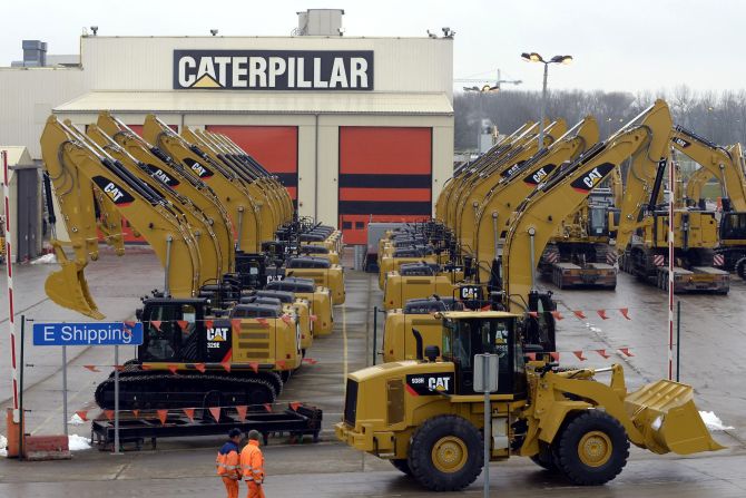 Workers walk past Caterpillar excavator machines at a factory.