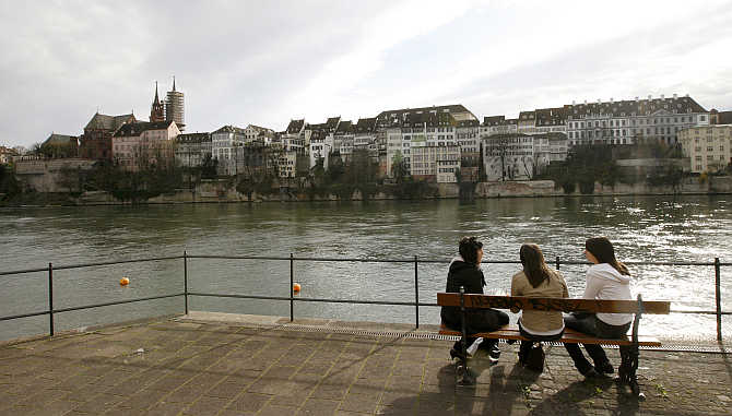 Girls sit on a bench on the banks of the Rhine River, in front of the old town of the northern Swiss city of Basel, Switzerland.
