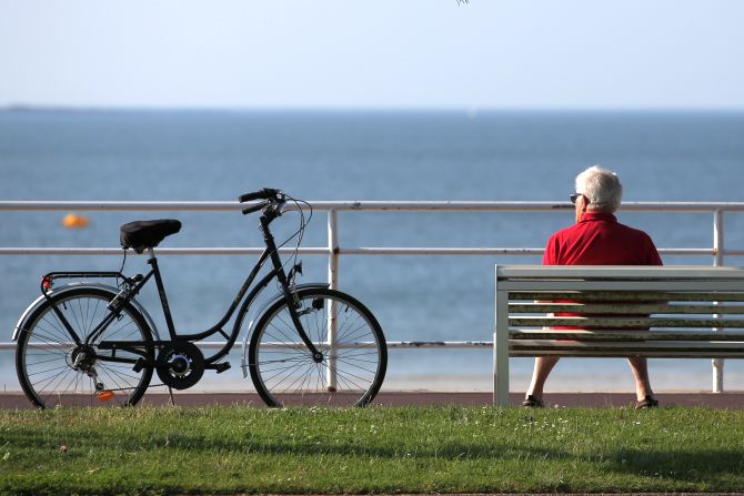 An elderly man sits on a bench next to his bicycle to take in the sun.