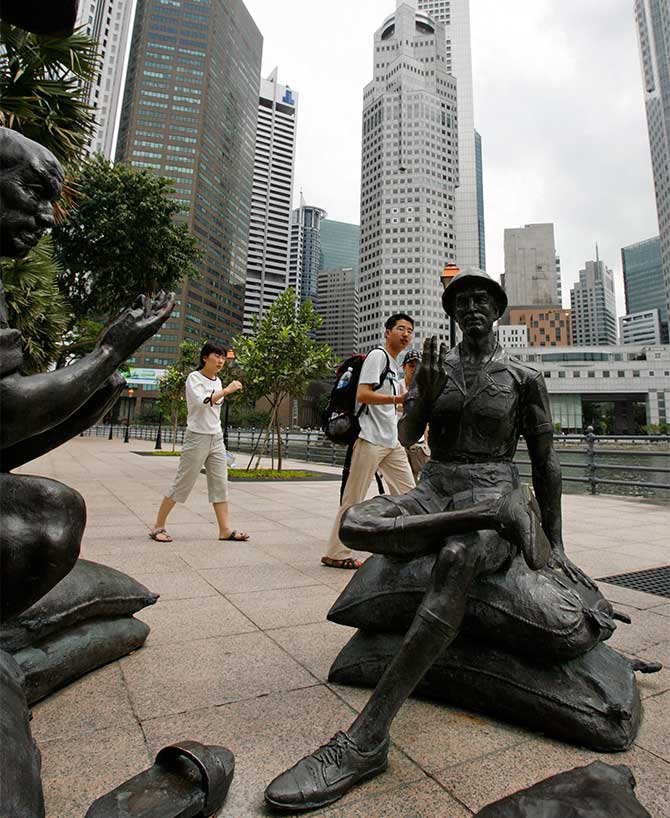 Tourists walk past statues along the Singapore river, with the financial district in the background.
