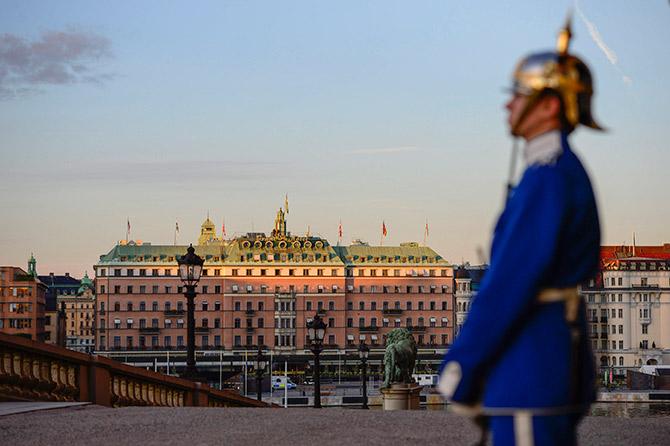 A palace guard stands in front of the Royal Palace while the sun sets over the Grand Hotel in downtown Stockholm.