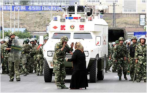 A woman on a crutch argues with a Chinese soldier in front of an armoured personnel carrier and soldiers wearing riot gear