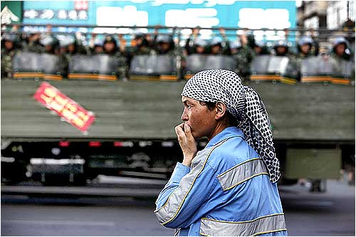 An ethnic Uighur woman stands on the side of the road as Chinese troops ride on a truck